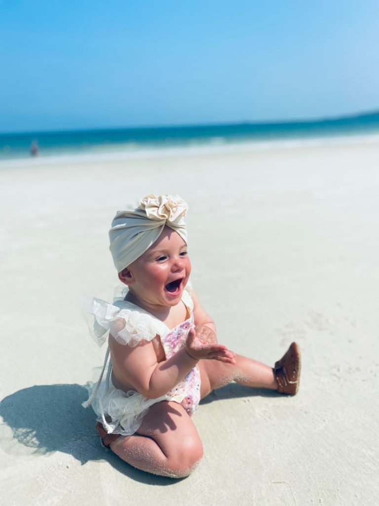 Een schattige baby op het strand, dragend een turban (mutsje), met een vrolijke uitdrukking op het gezicht.